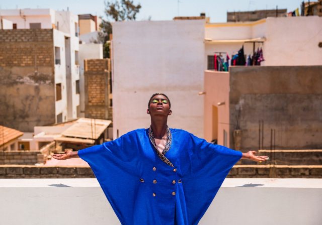 Eine Schwarze Frau im blauen Kleid streckt die Arme aus und guckt Richtung Himmel. Sie steht auf einem Dach.